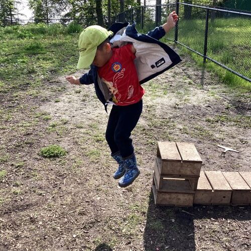 A preschool boy is mid-air jumping off a small wooden block with arms out like he is learning to fly