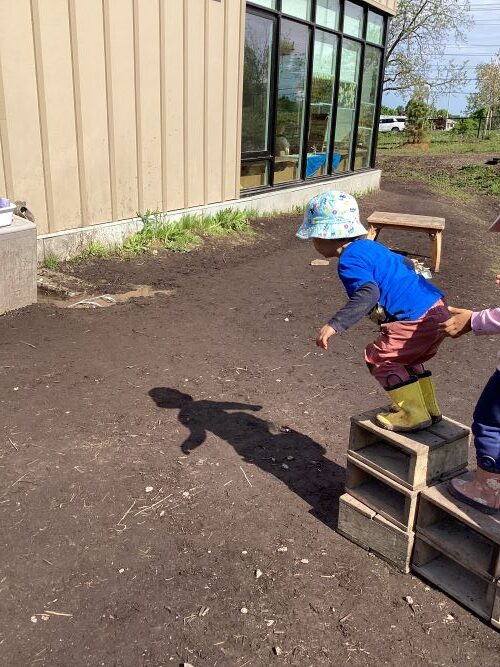 Boy with a bright blue shirt and yellow rain boots preparing to jump off a small wooden block.
