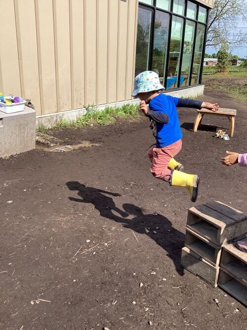 Boy with a bright blue shirt and yellow rain boots is mid-air jumping off a small wooden block.