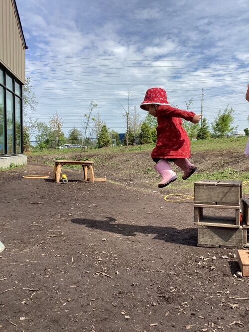 A preschool girl in red clothing is mid-air jumping off a small wood block onto the ground