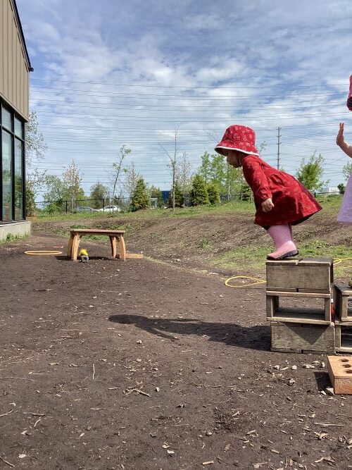A preschool girl in red clothing is about to jump off a small wood block onto the ground