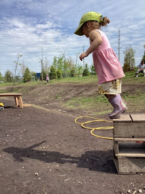 A preschool girl in a pink dress is about to jump off a small wood block onto the ground