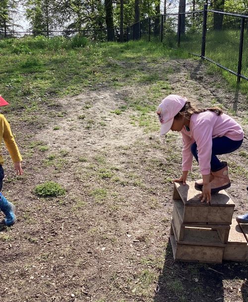 A preschool girl in a pink hat and shirt is climbing a small wood block