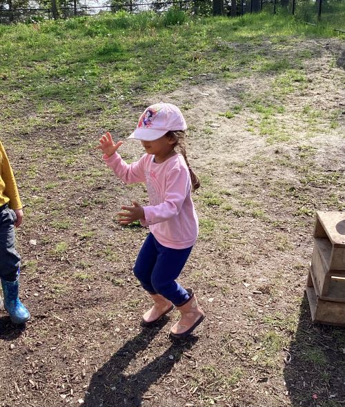 Girl in a pink hat and shirt has landed on the ground after jumping off a small wooden block.