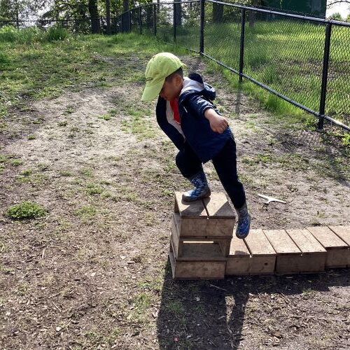 A preschool boy is climbing a small wooden block to jump with arms out like he is learning to fly