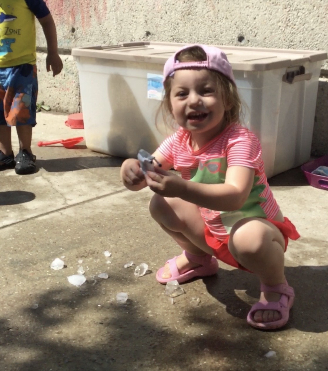 Smiling girl crouched on concrete, holding a chunk of ice with broken pieces scattered around.