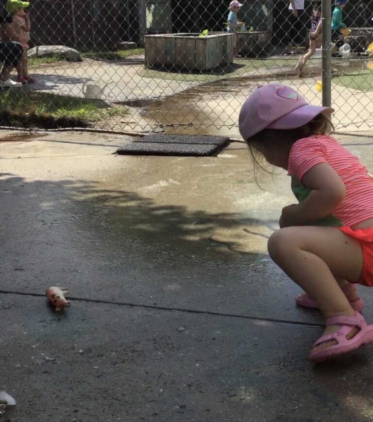 Child in pink swimsuit crouching on wet pavement, observing a toy animal and ice nearby.