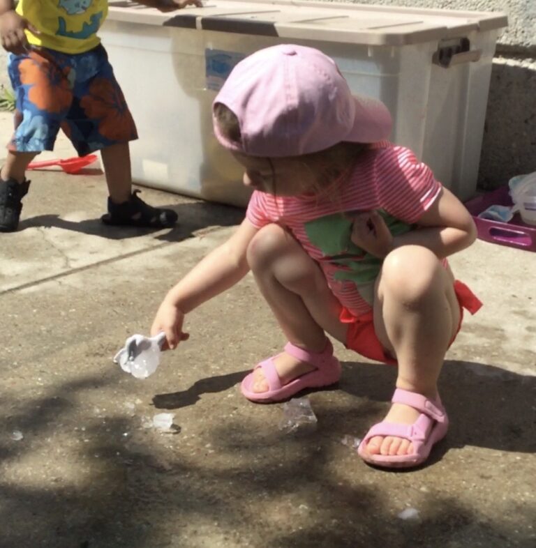 Child in pini outfit crouching on pavement while placing a block of ice on the ground.