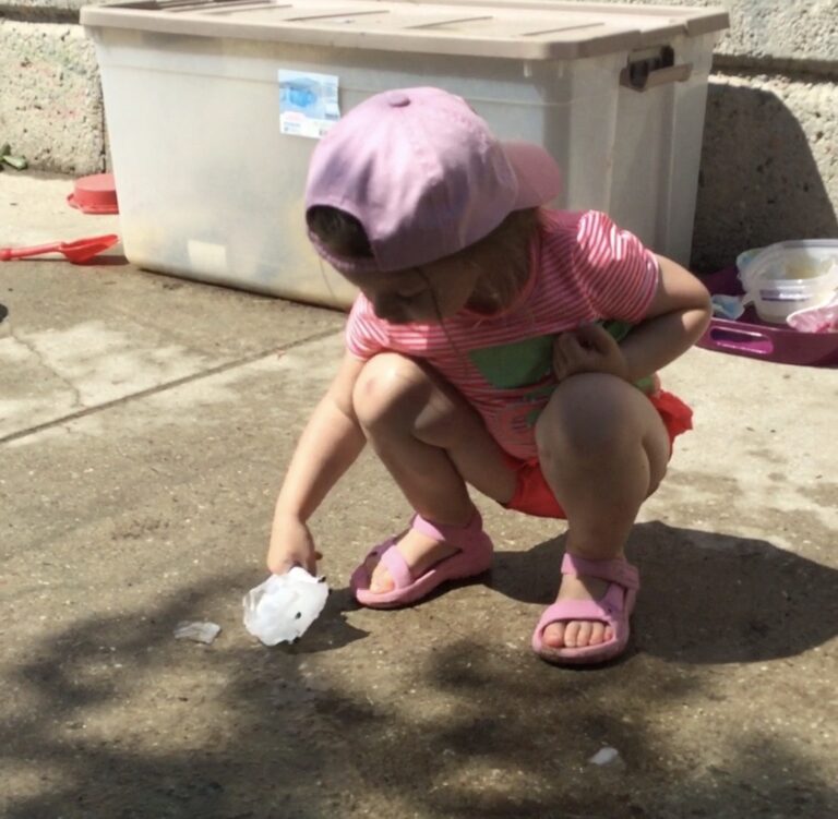 Child in pink summer clothes crouched, preparing to drop a block of ice onto the concrete.