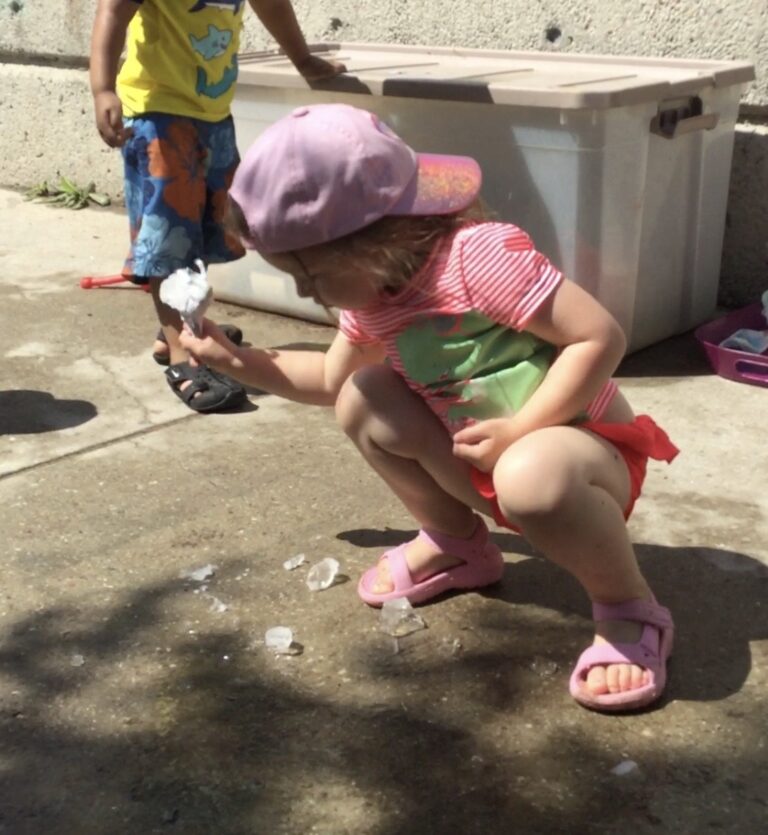 Child crouching, holding a melting chunk of ice over a pile of scattered ice pieces.