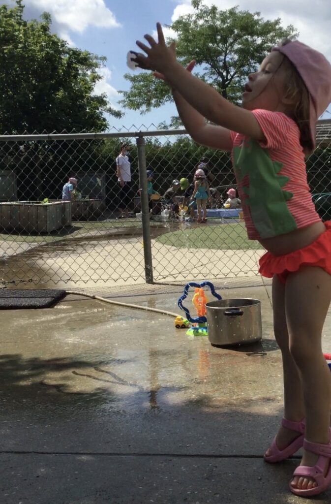 Child in pink swimsuit throwing ice upward with excitement during outdoor play.