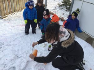 Educator kneeling in the snow assisting children with their coloured spray bottles.