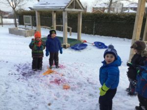 Children standing on a snow-covered playground admiring the colourful snow artwork they created.