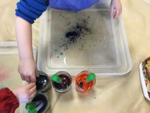 Close-up of children’s hands using droppers to mix different colours into jars and a water tray.