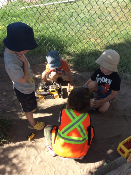 Four children playing with toy construction trucks in the sand, one child wearing reflective vest.