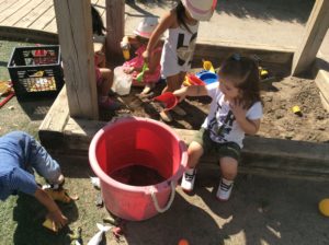 Several children playing with shovels, animals, and buckets in a sandbox under a wooden shelter.