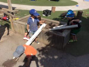 Three children use long plastic pipes to move soil from wooden planter box to ground.