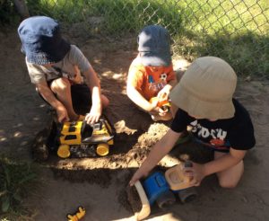 Three children playing with a construction vehicle in sand.