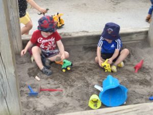 Two children sit in sandbox playing with toy dump trucks and sand toys.