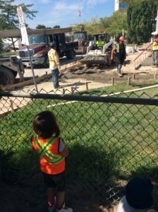 Child wearing safety vest stands at the fence, observing real construction.