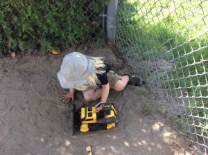 A preschooler in a wide-brimmed hat plays alone in a quiet corner with a large yellow toy bulldozer.