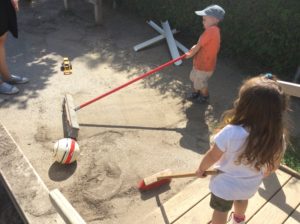 Two children using brooms to push sand and a toy construction truck outside in a sunny play area.