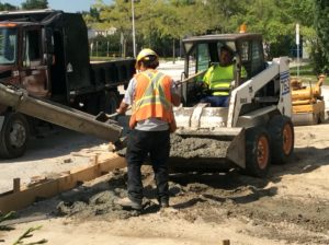 Worker guiding a cement pour from a truck into a wooden form while another operates the Bobcat.
