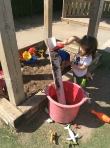 Child using a red shovel to scoop sand into white pipe in a pink bin outside.