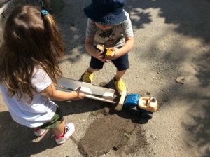 Two children pouring sand down a white pipe over a toy construction worker on the pavement.
