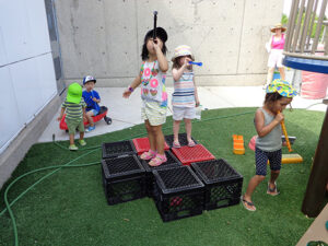 Children playing outdoors, standing on stacked milk crates and using toy telescopes.