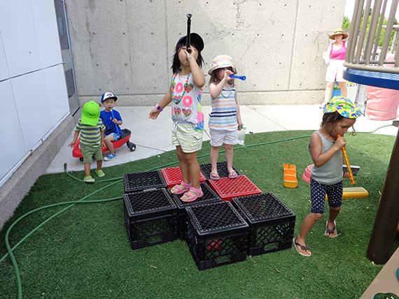 Children playing outdoors, standing on stacked milk crates and using toy telescopes.