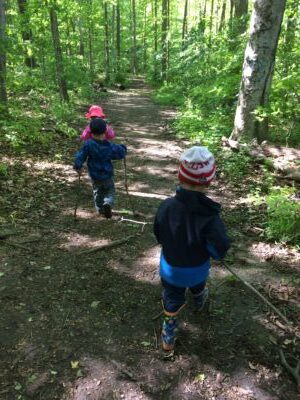 Toddlers walking down a forest path holding long sticks in their hands.