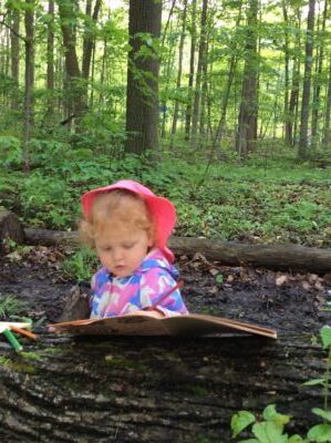 Girl in a pink sunhat reading a book on a fallen tree in the forest.
