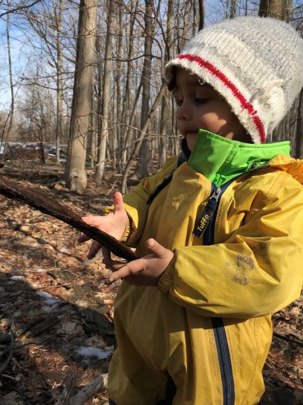 Child in a yellow rain suit and knit hat examining a large stick.