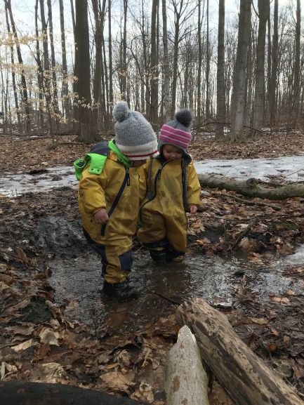 Two children standing on icey puddle surrounded by fallen leaves in forest.