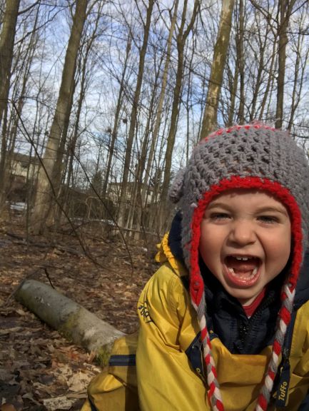 Smiling child in knit hat and yellow jacket laughing with joy in forest covered in dry leaves.