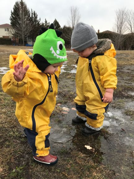 Two toddlers in yellow rain suits are looking at icy patch on the ground.