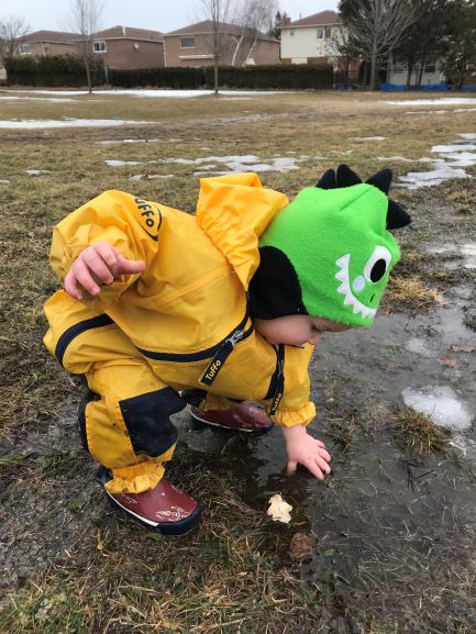 Child in a green monster hat and yellow rain suit reaching toward a leaf in puddle outdoors.