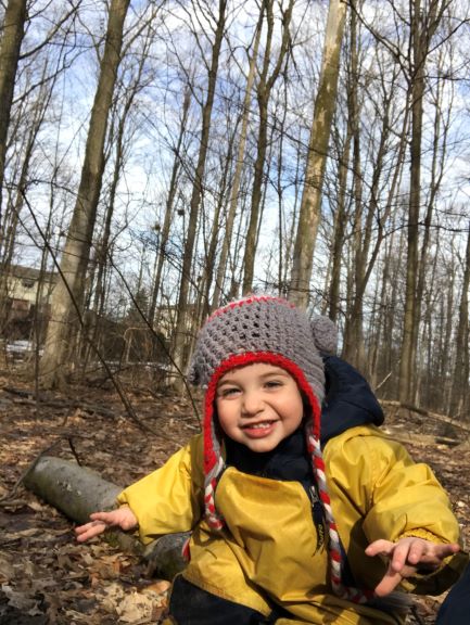 Smiling child in a yellow rain suit and hat in forest, surrounded by leaf-covered ground.