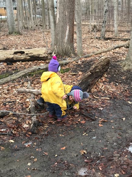 Two children wearing winter hats and rain suits kneeling beside a fallen tree trunk.