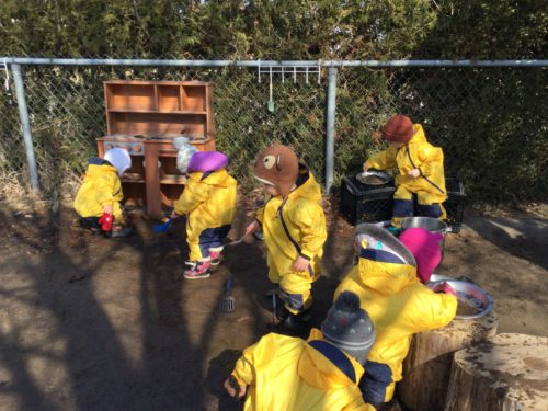 Children in yellow rain suits playing at an outdoor mud kitchen setup.