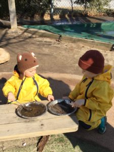 Toddlers sitting outdoors sharing pretend food made with mud.