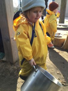Child carrying large pot filled with mud while exploring outdoors.
