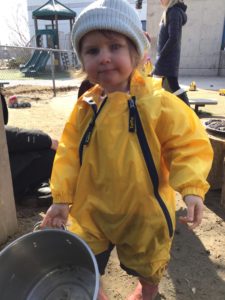 Toddler holding a pot and smiling during outdoor mud kitchen play.