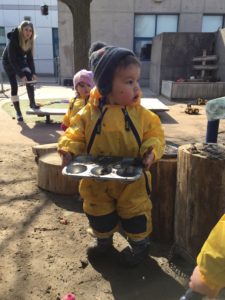 Child in yellow rain suit carrying tray filled with mud creations.