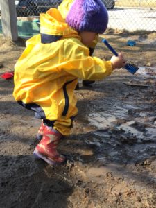 Child raking through a puddle with toy rake during mud exploration.