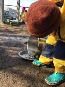 Child pouring mud from a pot into a metal dish during outdoor play.