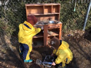 Children using kitchen tools and mud in outdoor play kitchen.