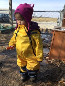 Toddler holding a whisk during muddy outdoor play in bright rain gear.