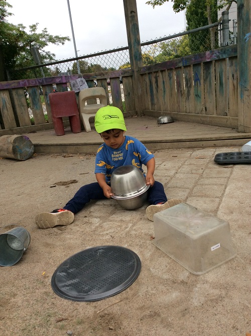 Child sitting on the ground outdoors, exploring sound of two metal bowls as improvised drums.